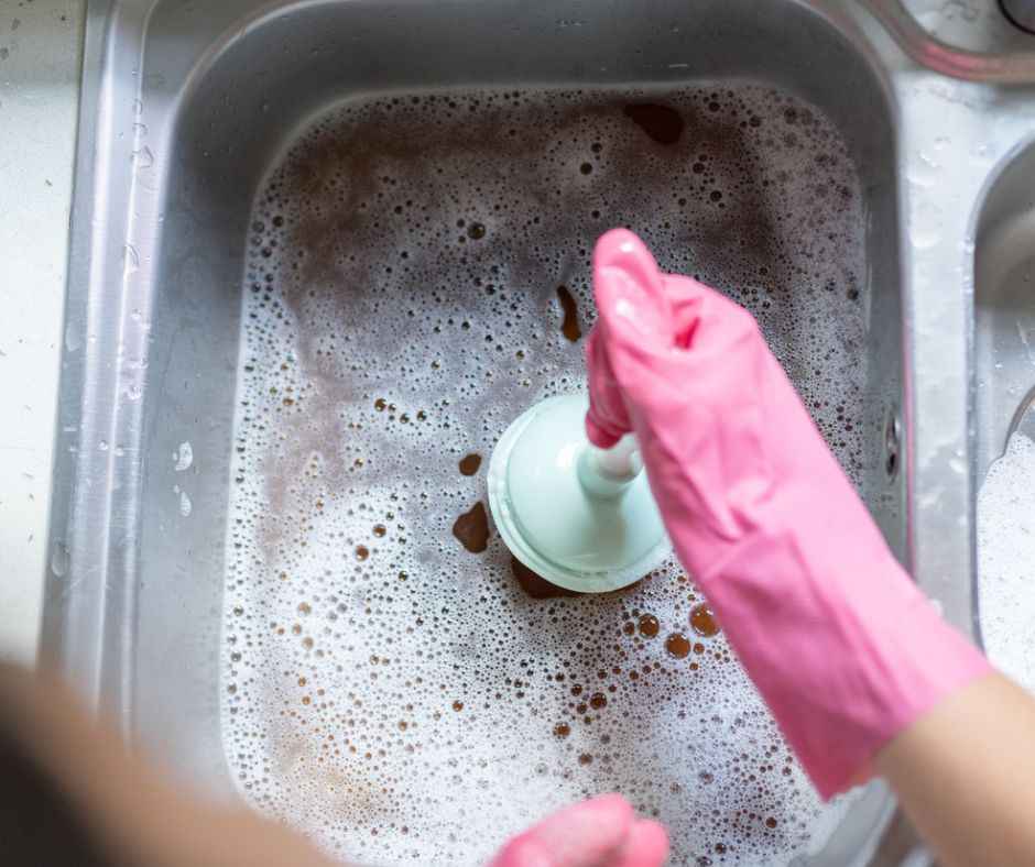 An adult woman using a plunger to remove a kitchen sink drain clog while wearing pink rubber gloves.